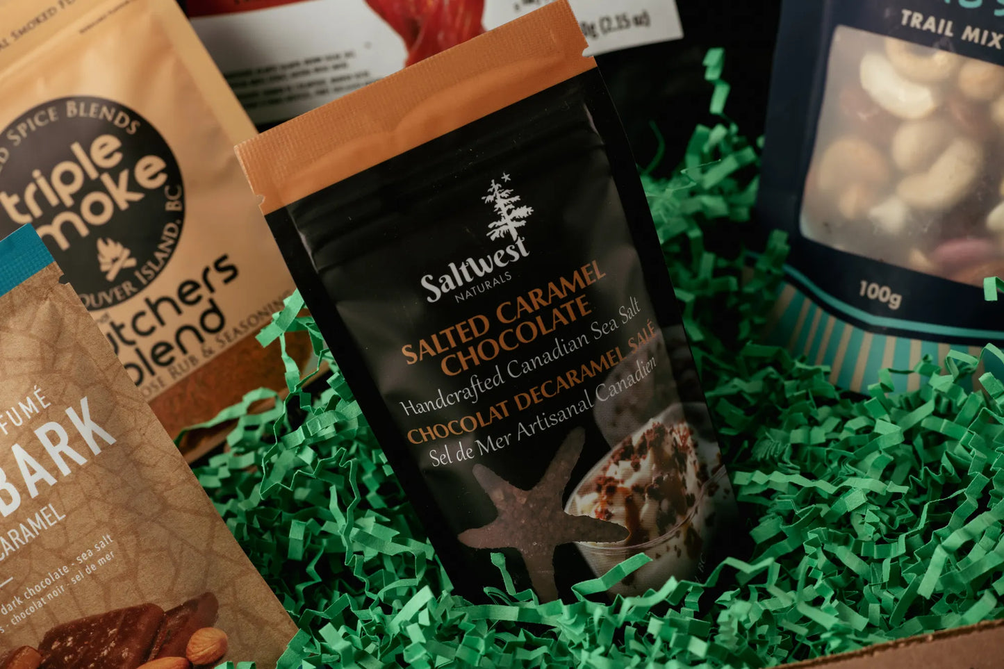 Assorted snack packages including Saltwest Salted Caramel Chocolate and Triple Moke Dark Chocolate Bark on green shredded paper.